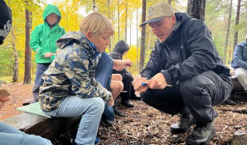 Pedagog visar elev hur man täljer en pinne ute i skogen.