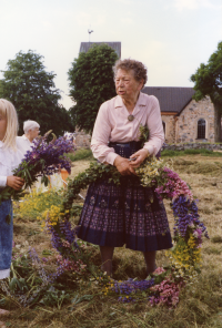 Midsommarstången på Kyrkängen i Vallentuna förbereds kvällen innan midsommarafton. Anna-Brita Dahl binder en blomsterkrans som bland annat innehåller blå och rosa lupiner. Vallentuna kyrka med kyrktuppen på taket syns i bakgrunden.