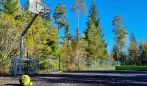 En asfalterad basketplan med en basketkorg till vänster i bild. På marken syns en basketboll och en tennisboll.