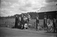 Folksamling runt mjölkpallen vid vägskälet utanför Lillgården i Kårsta. Från vänster Hugo Nyberg, Karl-Erik Nyberg och Birger Nyberg (svensk mästare i tresteg). Ståendes på mjölkpallen Gunilla Bergström. Sittandes överst Åsa Ahlgren och Lillemor. Sittandes nedre raden Barbro Bergström och Irene Lundin. Timo Ekholm står till höger om mjölkpallen, hans cykel ligger i diket till vänster. Namn saknas på pojken längst till höger.