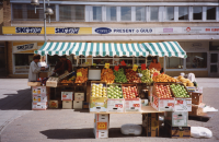 Torghandel med frukt och grönt på Tuna torg. I bakgrunden syns Torghusets fasad med skyltar för skoaffär, present- och guldaffär, frisör och elbutik.