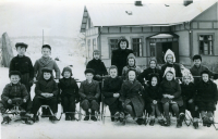 Lärarinnan Gunborg Sjöström fotograferas utanför skolan tillsammans med barnen i Össeby-Garns folkskola. Birgitta Lindell klass 2 sitter på sparken trea från höger. 
Namn textat på baksidan, från vänster bakre raden,  Yngve A, Lars R, Arne J, fröken Sjöström, Gunilla E, Britt A, Ragna A och S Hägg?. 
Främre raden, Olle P, Göran ?, Lars G, Leif J, Ing? K, Eivor J, Margareta B, Birgitta Lindell, Britt N och Hans P. Barnen är vinterklädda och raden längst fram sitter på sparkstöttingar och kälkar. Snö täcker skolgården och skolhusets tak. Årtal saknas.