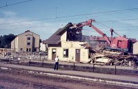 Rivning av Vallentuna stationshus pågår. En grävskopa hugger tag i resterna av stationshuset. I bakgrunden till vänster syns flerfamiljshus vid Centralvägen.